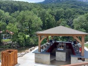 Un espace extérieur abrité, doté d’une structure en bois, d’un comptoir en béton et offrant une vue sur une rivière et des collines boisées.