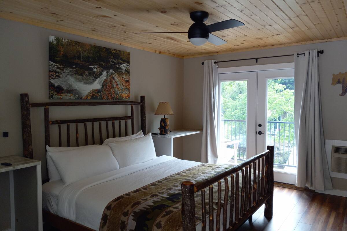 Ein Schlafzimmer mit einem hölzernen Bett, Deckenventilator, einem Bild eines Wasserfalls, einem Nachttisch, einer Lampe und einem Balkon mit Vorhängen.