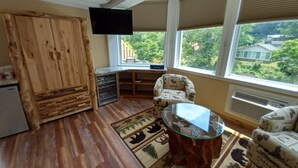 A cozy room with a wooden floor, a glass table, two patterned chairs, a TV, and large windows with a view of greenery. - The Carter Lodge On The River  (Chimney Rock)