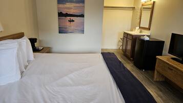 A hotel room with a bed, a painting of a sunset over water, a desk with a lamp, a dresser, and a television.