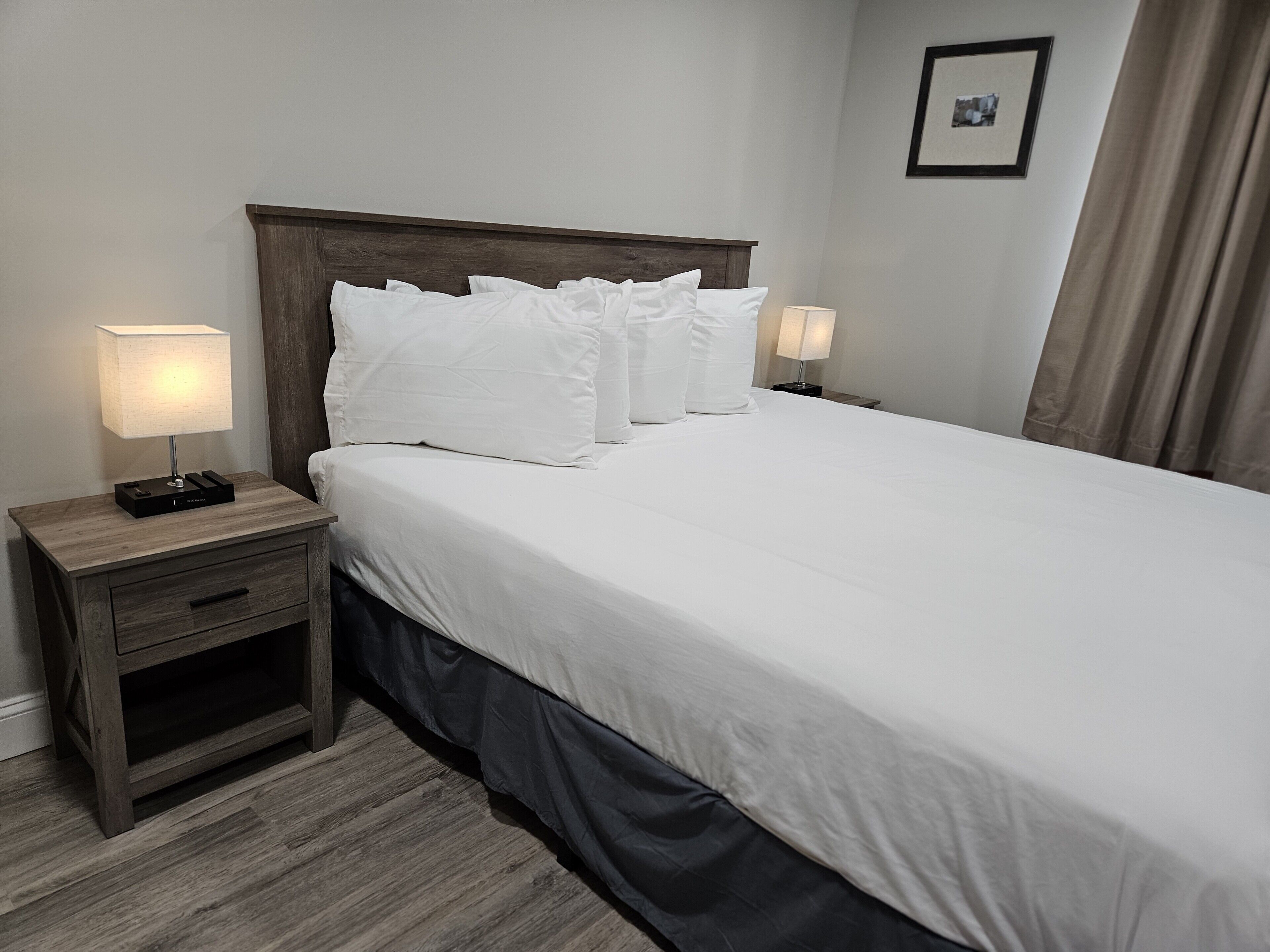 Een opgemaakt bed met witte lakens en kussens, een houten nachtkastje met een lamp, en een lijst met een foto aan de muur.
