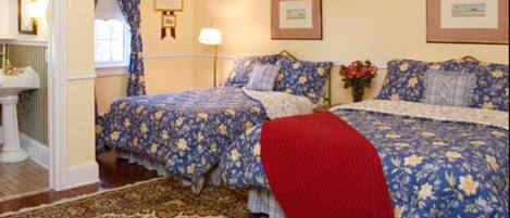 A bedroom with two beds, floral bedding, a red throw blanket, a nightstand, a lamp, and a framed picture on the wall.