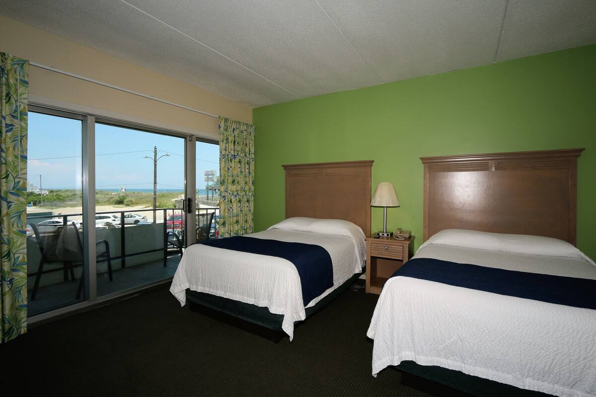 Ein Hotelzimmer mit zwei Betten, Blick durch ein großes Fenster und einem Balkon mit Stühlen.