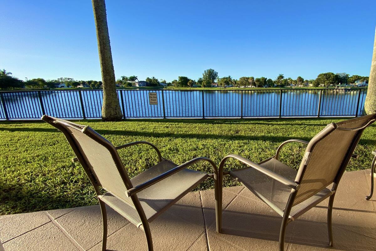 A patio area with two wicker chairs and a small table, overlooking a body of water and a fence.