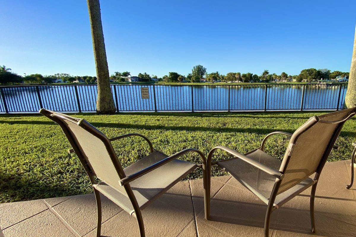 A patio area with two wicker chairs and a small table, overlooking a body of water and a fence.