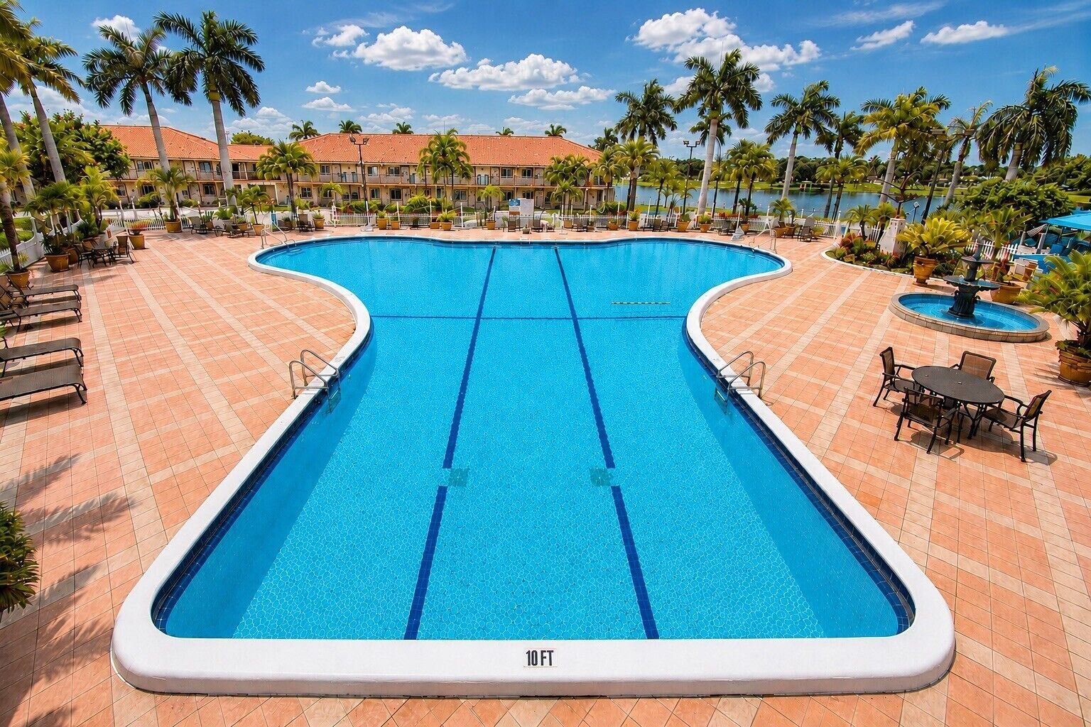 A large, clear blue swimming pool with a 10 FT depth marker, surrounded by lounge chairs and palm trees, with a building in the background.