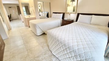 Habitación de hotel con una cama grande, dos camas más pequeñas, una mesita de noche con lámpara, un espejo y un baño al fondo.