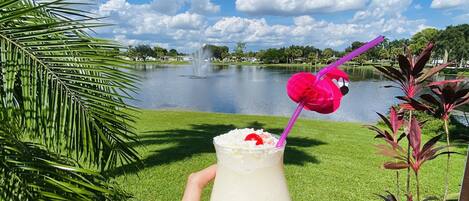 A hand holding a cocktail with a pink flamingo straw, palm leaves, and a lake view in the background.