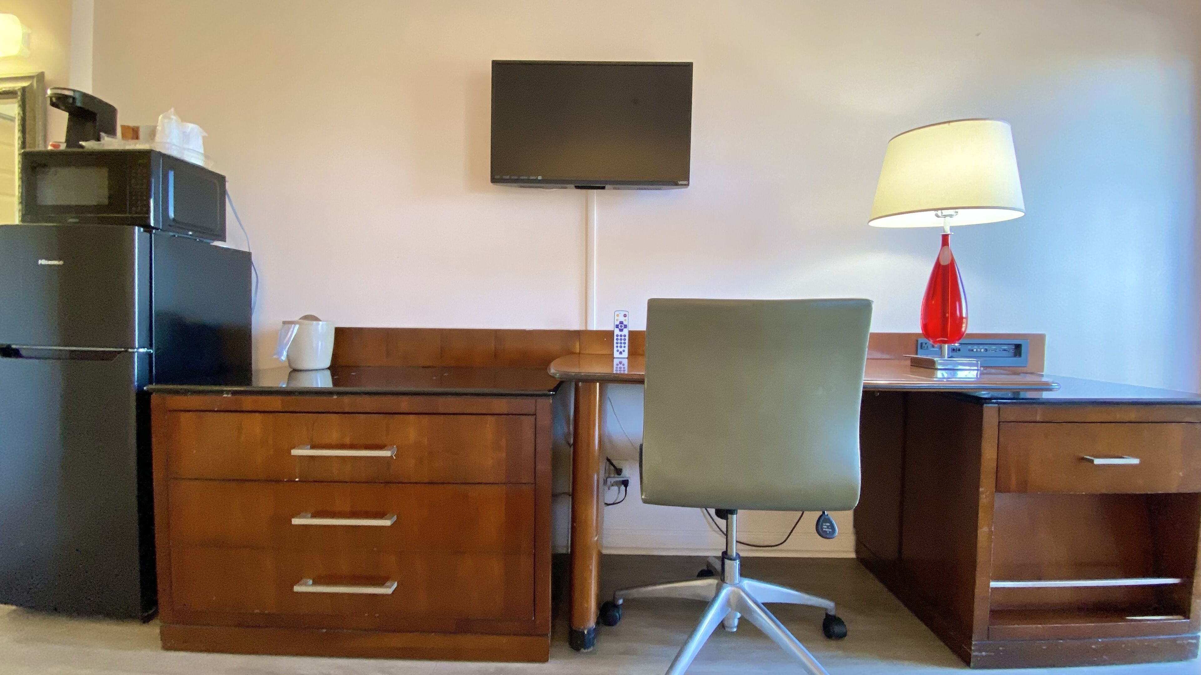 Ein Hotelzimmer mit einem Holztisch, einem Stuhl, einem an der Wand montierten Fernseher, einer roten Lampe und einem Kühlschrank.
