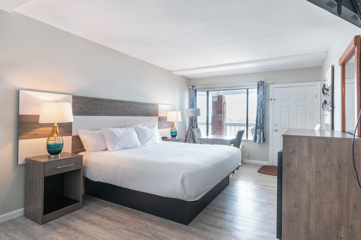 A modern hotel room with a large bed, bedside tables with lamps, a desk, and a view of the city through the window.
