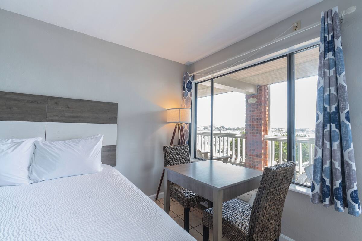 A hotel room with a large bed, a dining table, wicker chairs, a balcony with a view, and a patterned curtain.