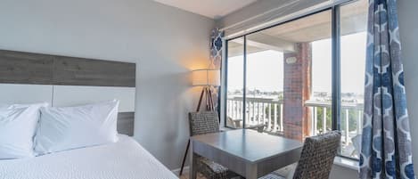 Una habitación de hotel con una cama grande, una mesa de comedor, sillas de mimbre, un balcón con vistas y una cortina estampada.