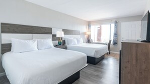 A hotel room with two beds, a wooden headboard, a nightstand with a lamp, a desk with a television, and a window with curtains. - The Anchorage Inn and Marina (Ocracoke)
