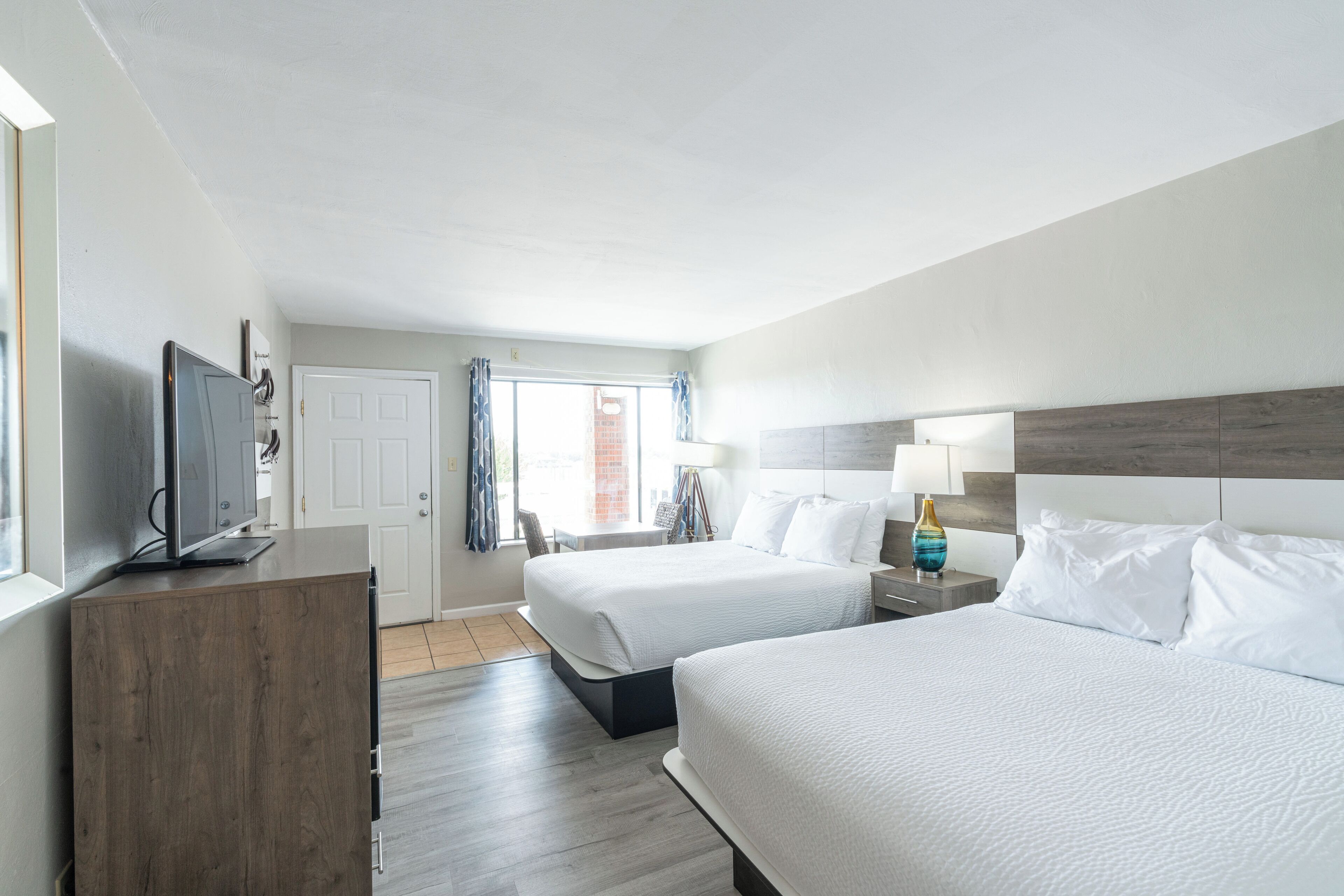 Habitación de hotel con dos camas, un televisor sobre un mueble de madera, una ventana con cortinas y una mesita de noche con lámpara.
