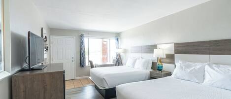 Habitación de hotel con dos camas, un televisor sobre un mueble de madera, una ventana con cortinas y una mesita de noche con lámpara.