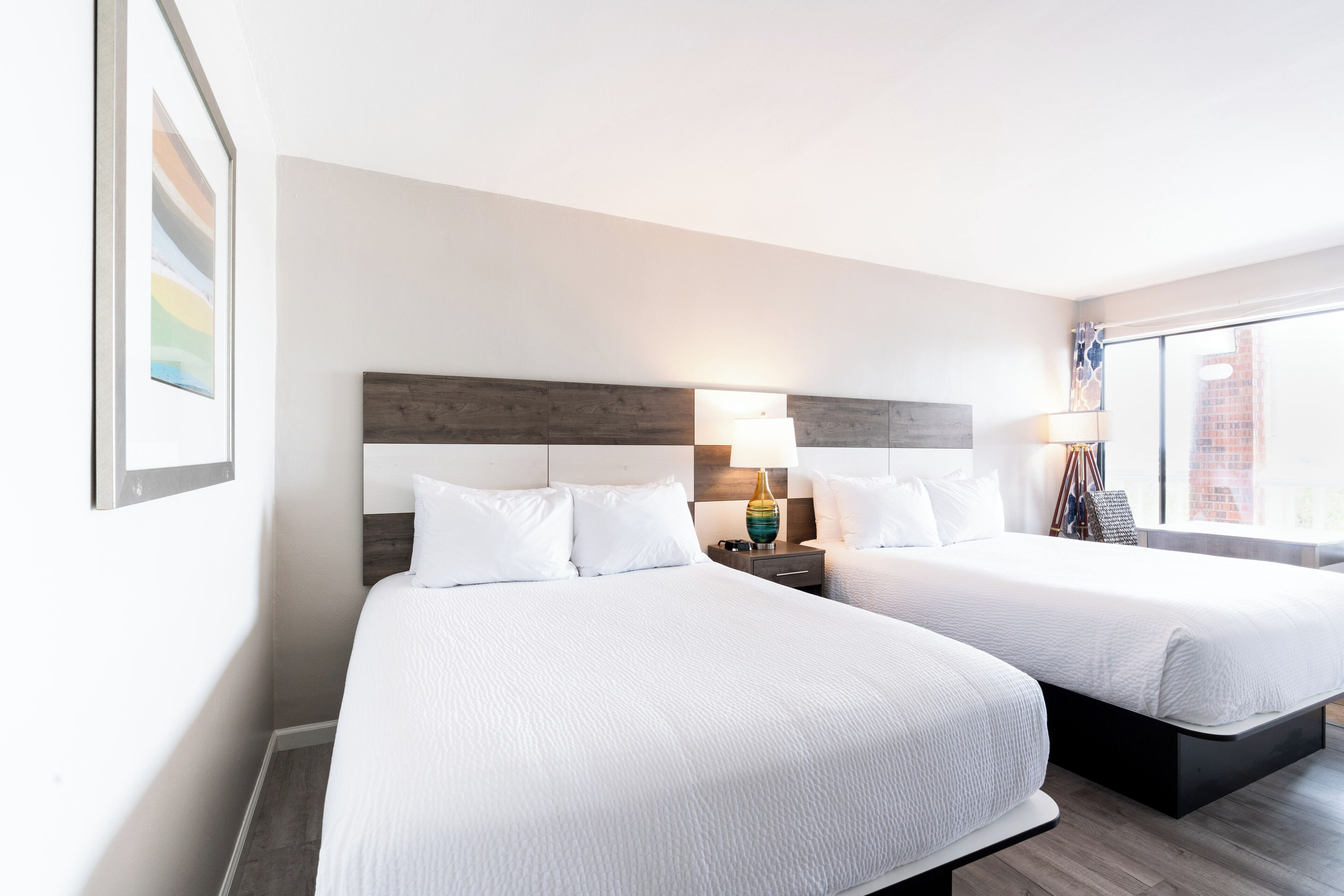 A hotel room with two beds, a nightstand with a lamp, and a window with a view of a building.