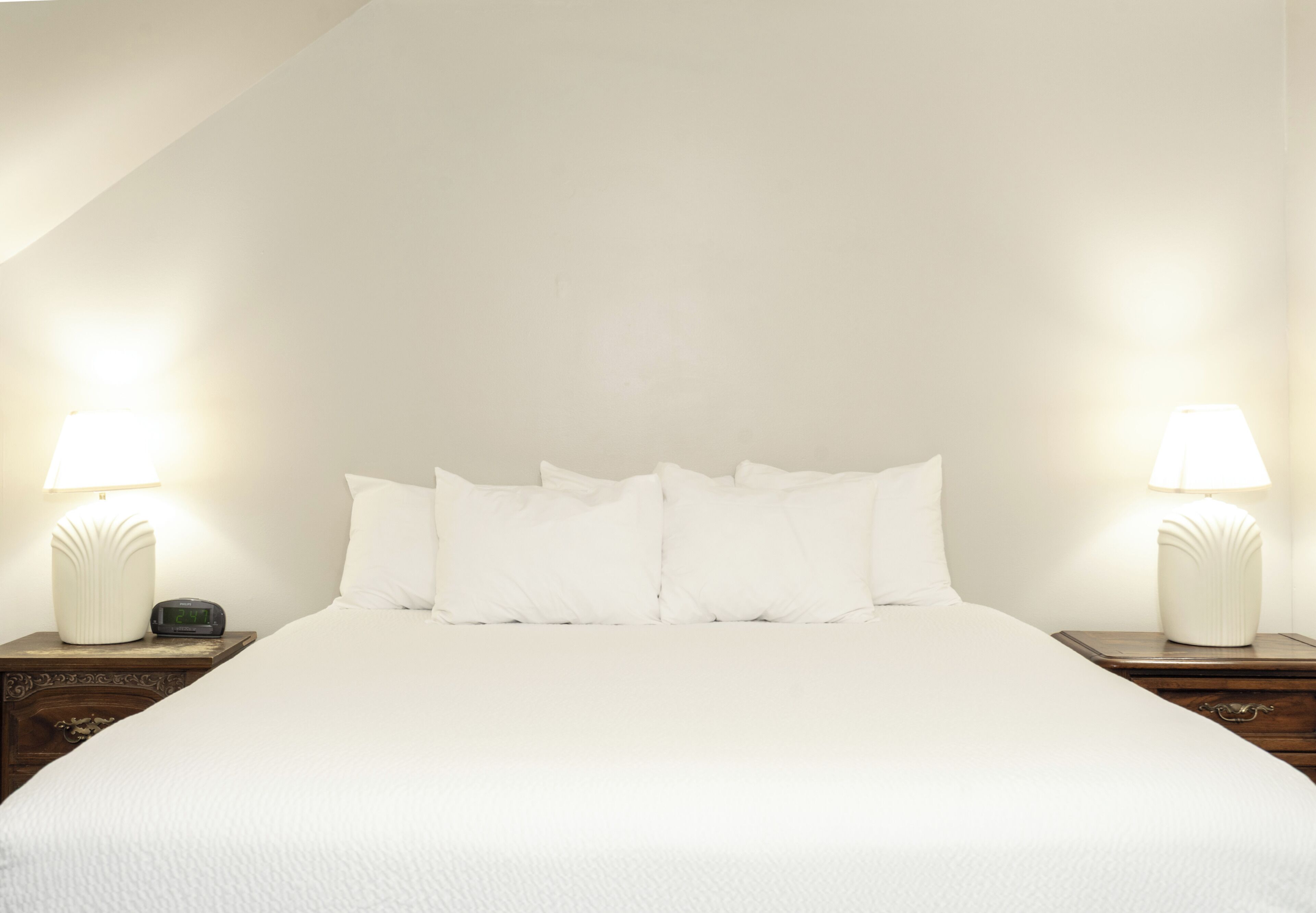 A neatly made bed with white linens and pillows, flanked by two bedside tables with lamps, in a room with plain walls and a sloped ceiling.