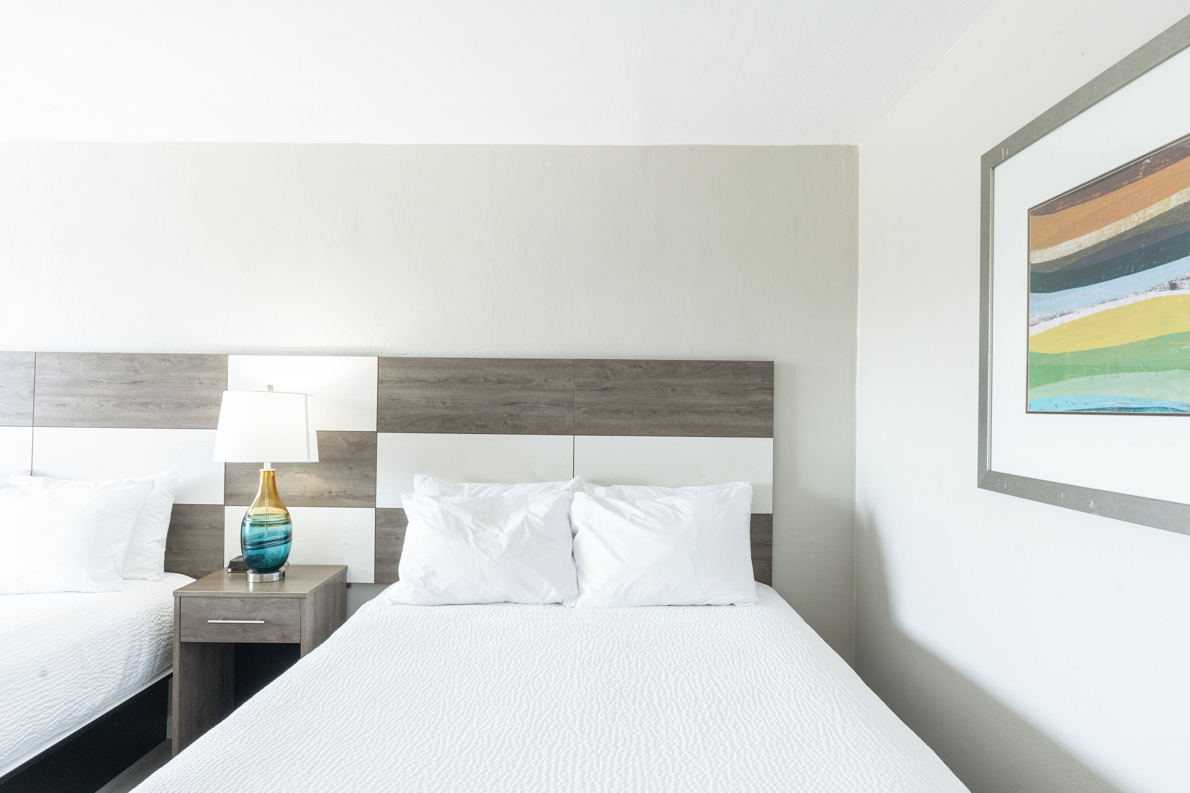 Habitación de hotel con dos camas, una mesita de noche con lámpara y un cuadro en la pared.