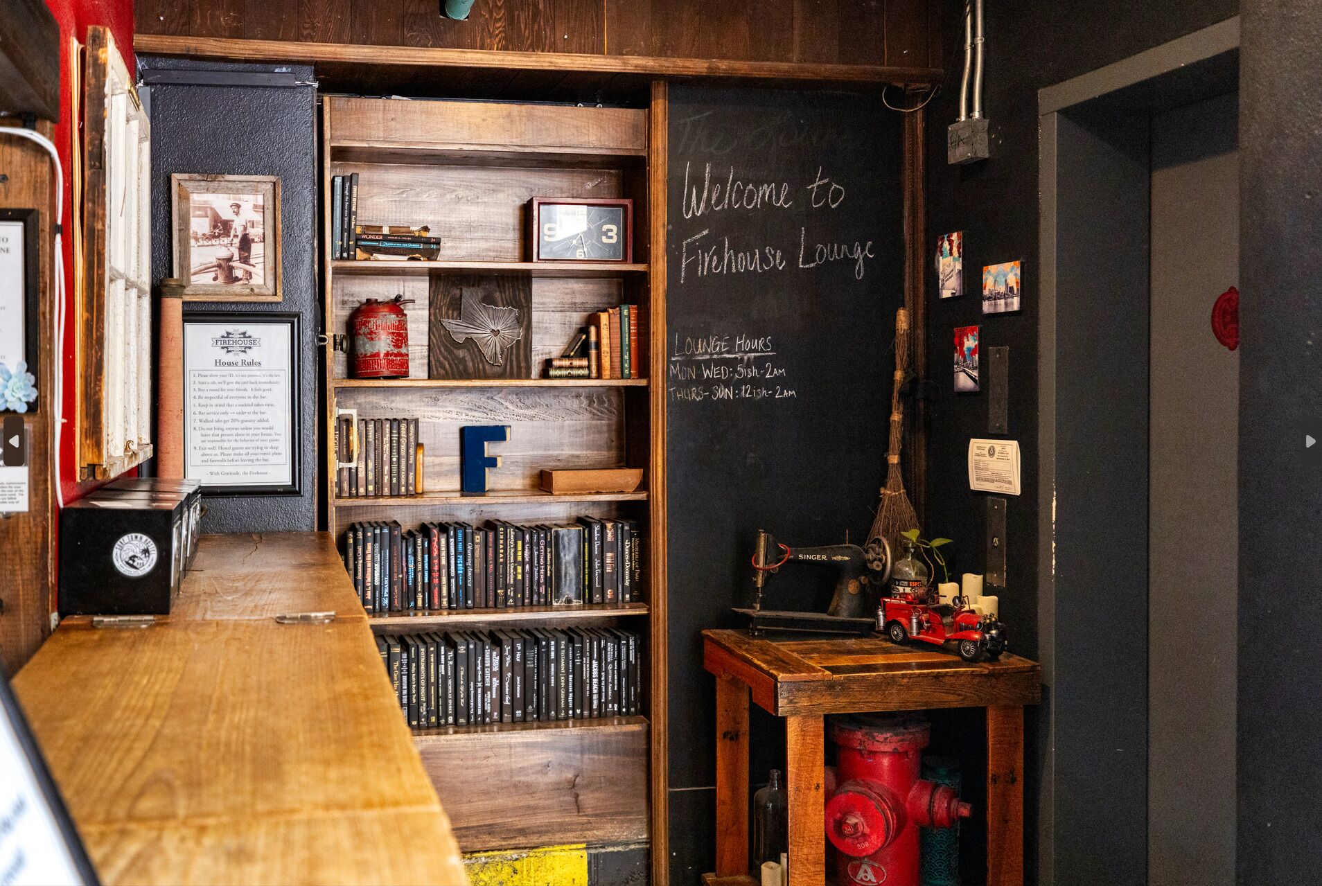 Un espacio de sala acogedor con una estantería de madera llena de libros, una pizarra con el horario de la sala y decoración vintage que incluye un hidrante y una máquina de coser Singer.