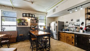 A kitchen with a wooden table and bar stools, stainless steel appliances, and a brick wall with clocks. - Firehouse Hostel (Austin)