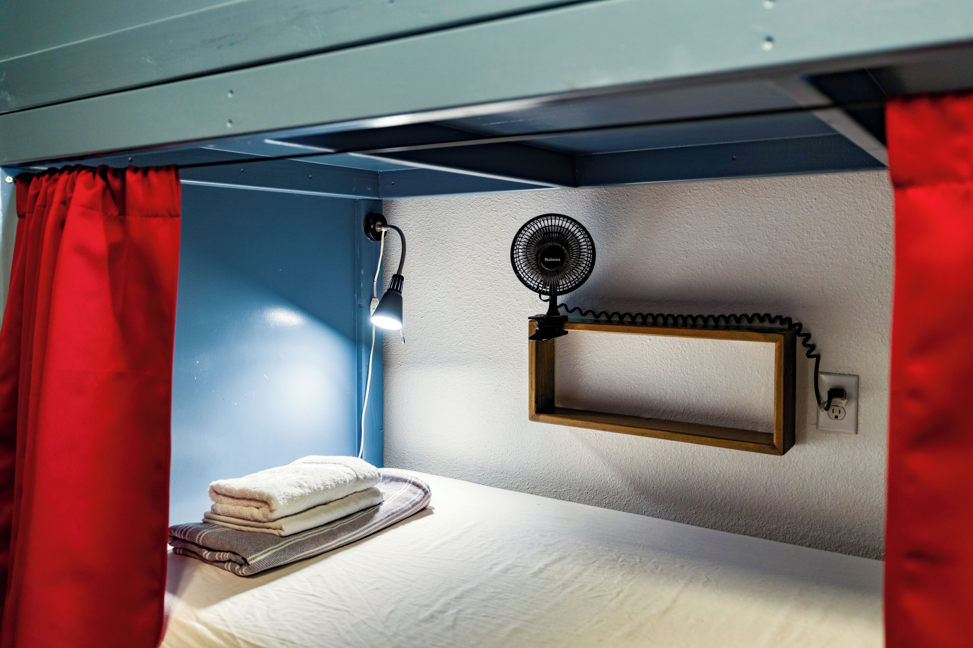 A bunk bed with a fan and a shelf, red curtains, and a bedside lamp.