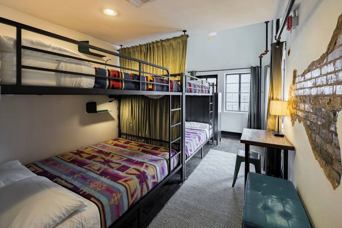 A bunk bed room with a desk and chair, a window with curtains, and a brick wall.