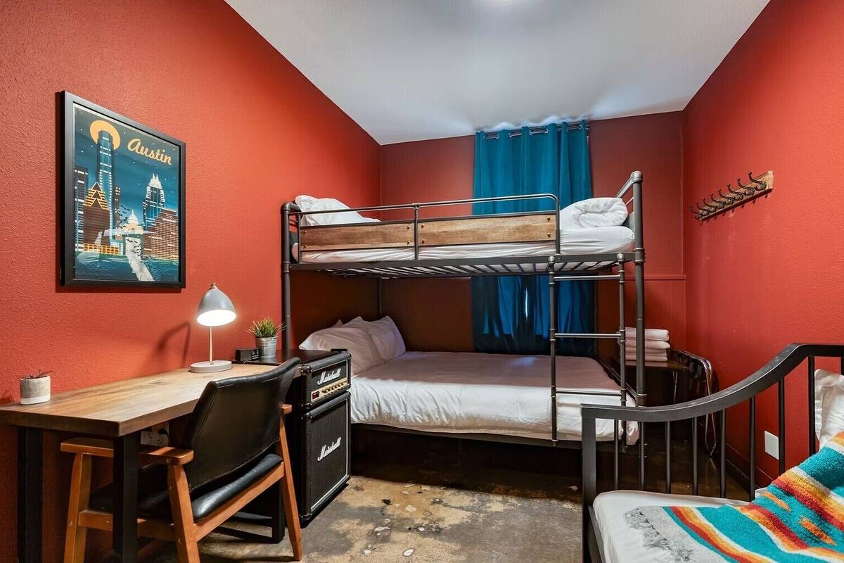 A dorm-style room with bunk beds, a desk with a lamp, a chair, a small table, and a colorful blanket.