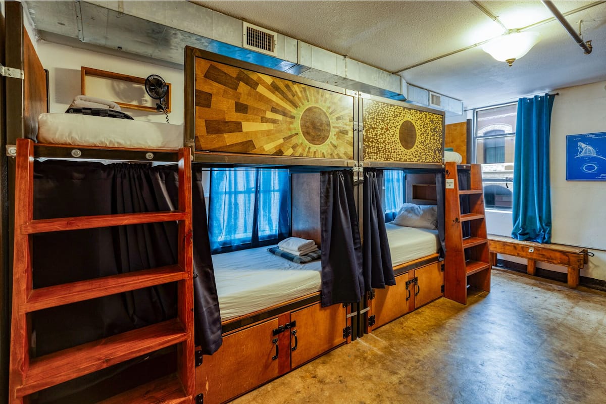 A dormitory-style room with bunk beds, a small desk, and a window with curtains.