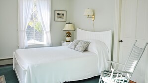 A bedroom with a bed, a nightstand with a lamp, a chair, a door, and a window with curtains. - The Claremont Hotel (Southwest Harbor)