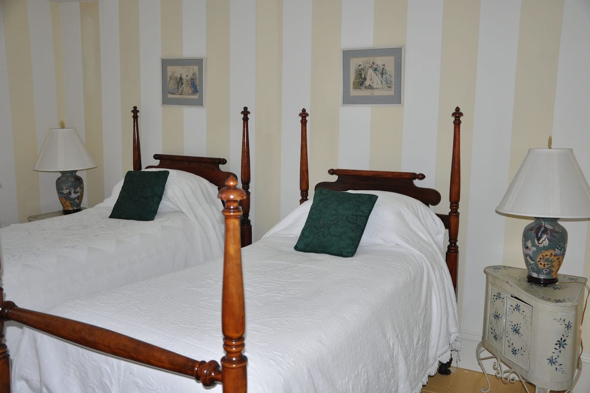 Ein Doppelbett mit Holz-Kopfteilen und grünen Kissen, ein Nachttisch mit Lampe sowie rahmengenannte Bilder an einer gestreiften Wand.