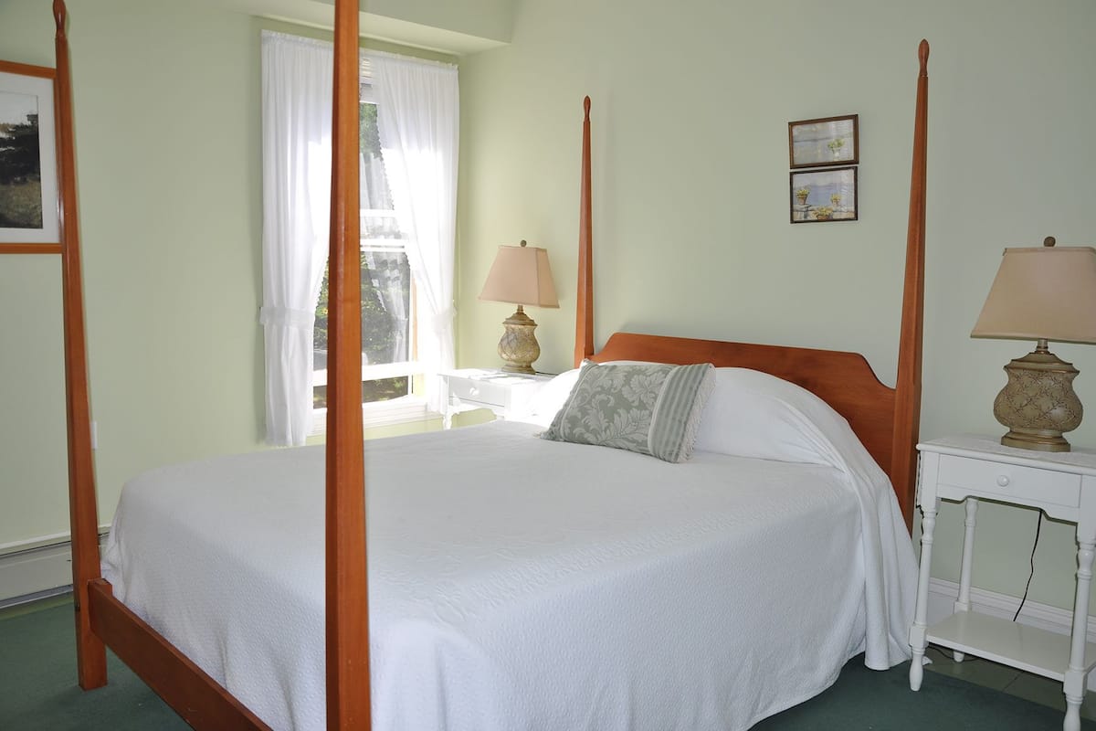 Ein Himmelbett mit weißer Bettwäsche, ein Nachttisch mit Lampe und ein Fenster mit weißen Vorhängen.