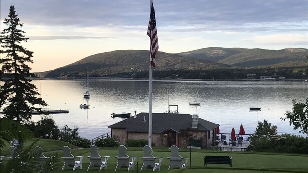 A flagpole with an American flag, a building with a roof, and a body of water with boats. - The Claremont Hotel (Southwest Harbor)