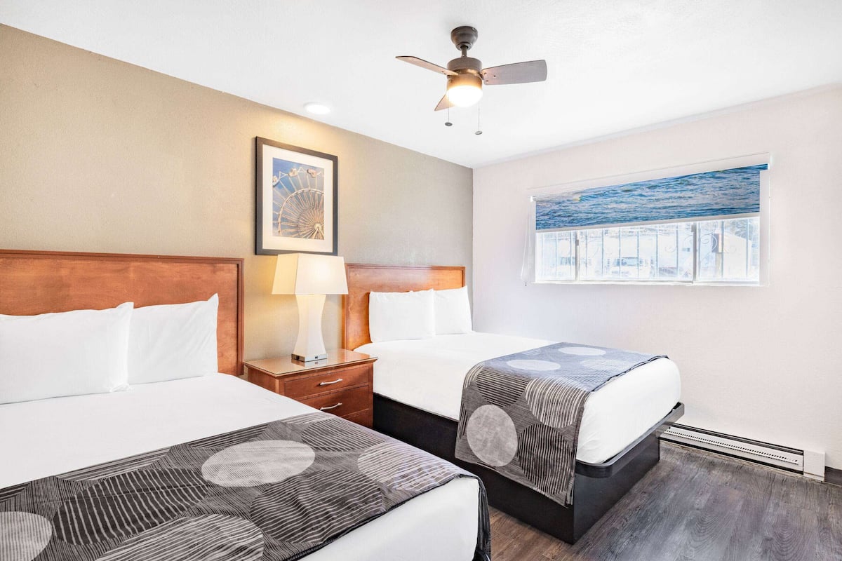 A hotel room with two beds, a ceiling fan, a nightstand with a lamp, a framed picture on the wall, and a window with a view of the sea.