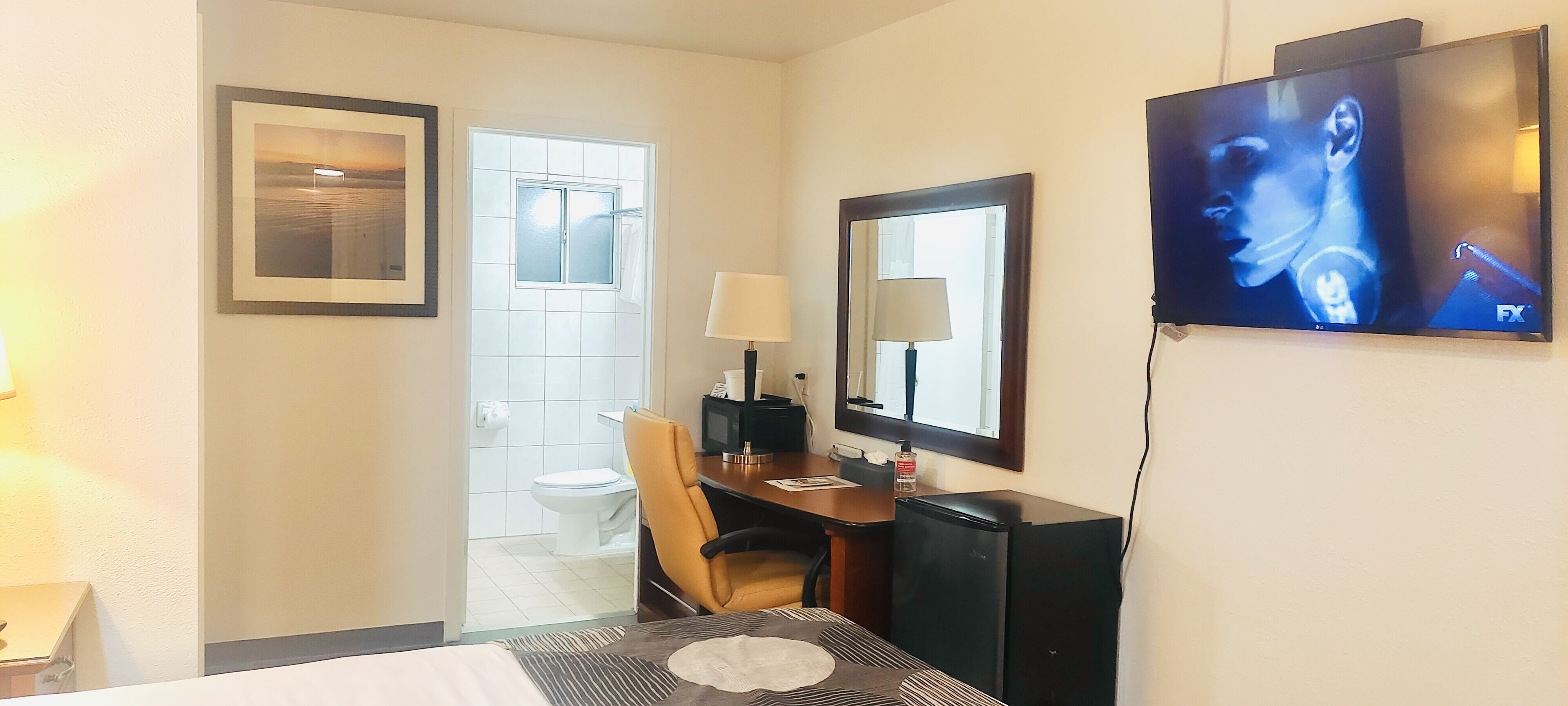 A hotel room with a bed, a desk with a chair, a television, a mirror, and a bathroom visible through an open door.