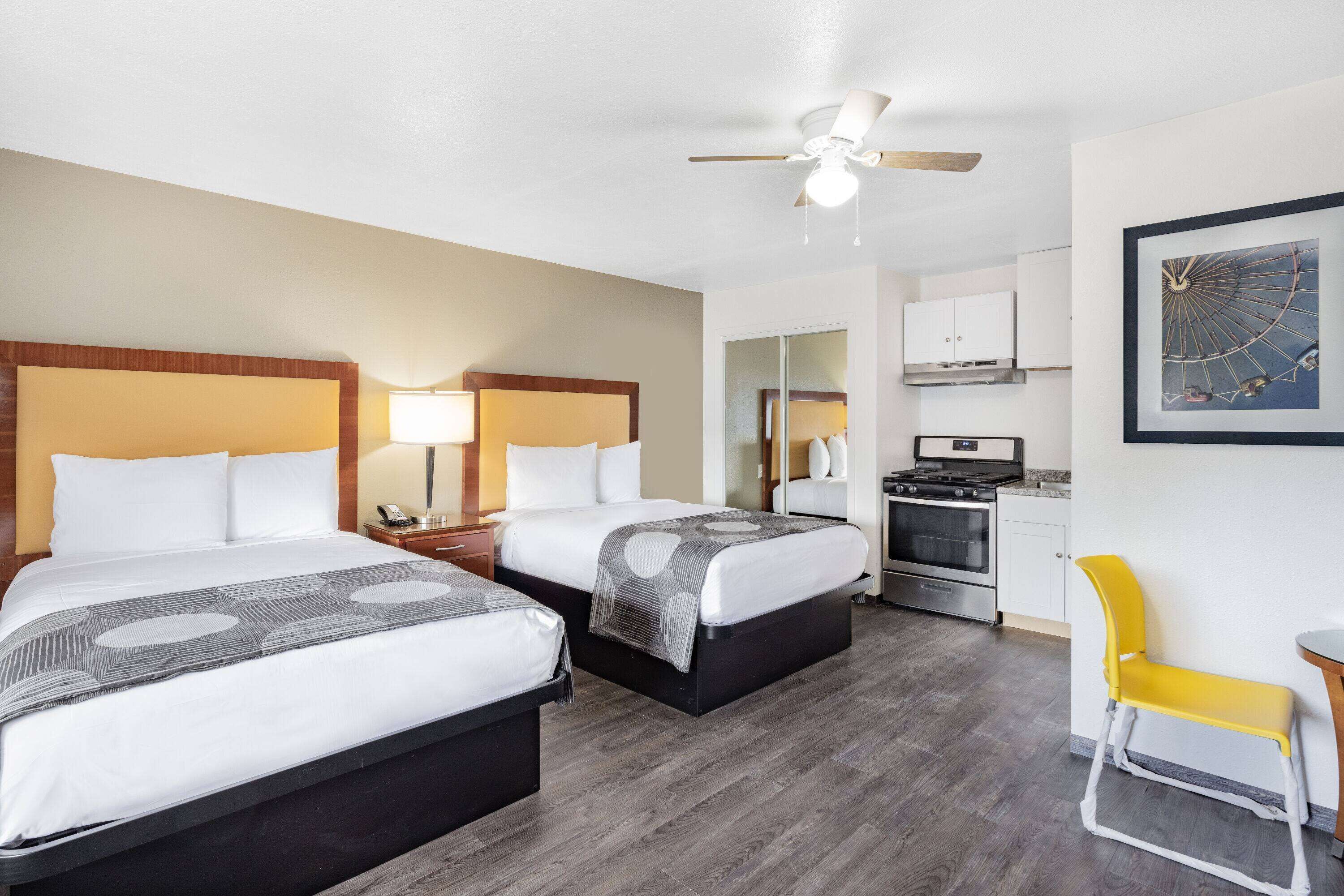 A hotel room with two beds, a desk, a chair, a kitchenette, and a ceiling fan.