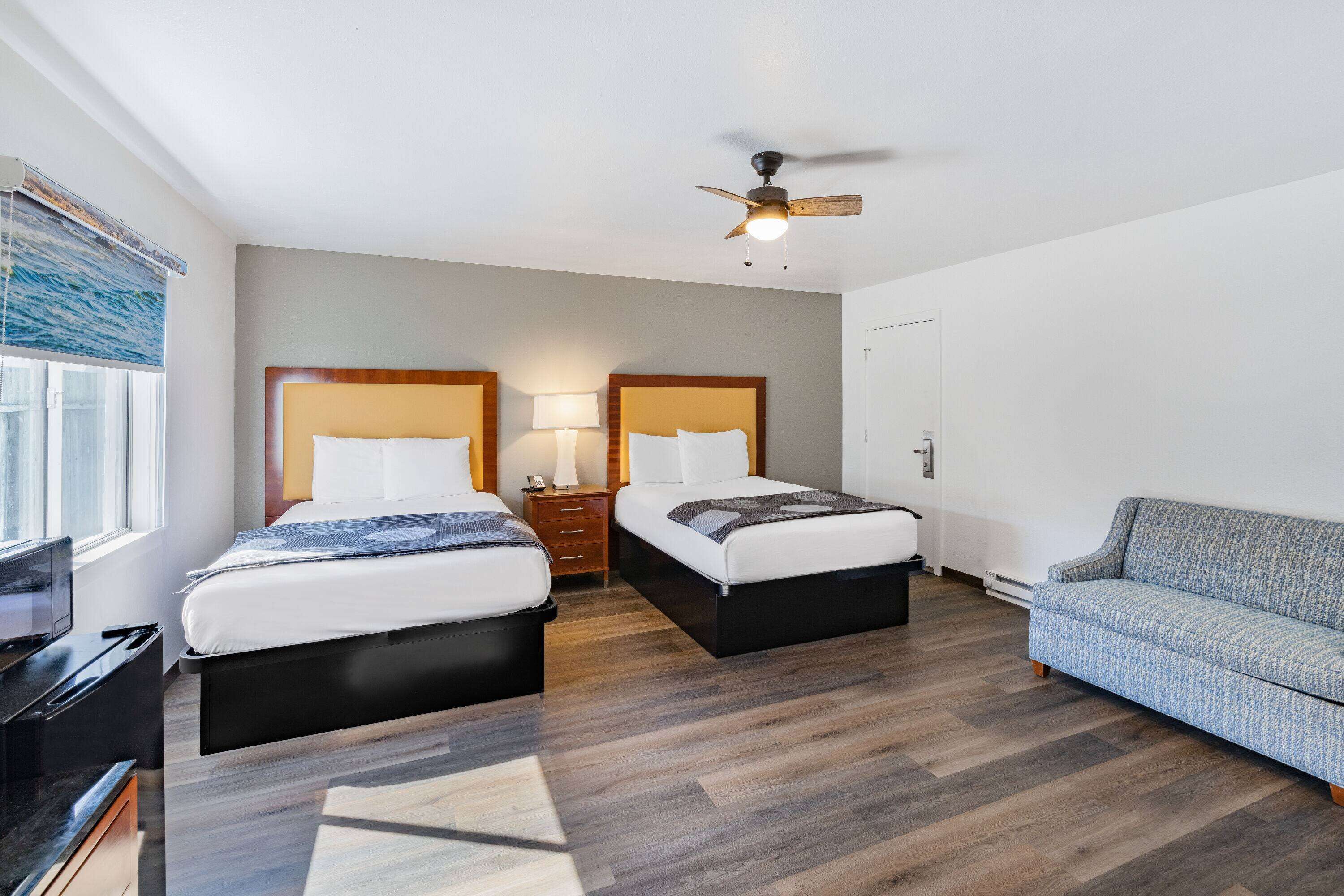 A hotel room with two beds, a sofa, a ceiling fan, and a window with blinds.