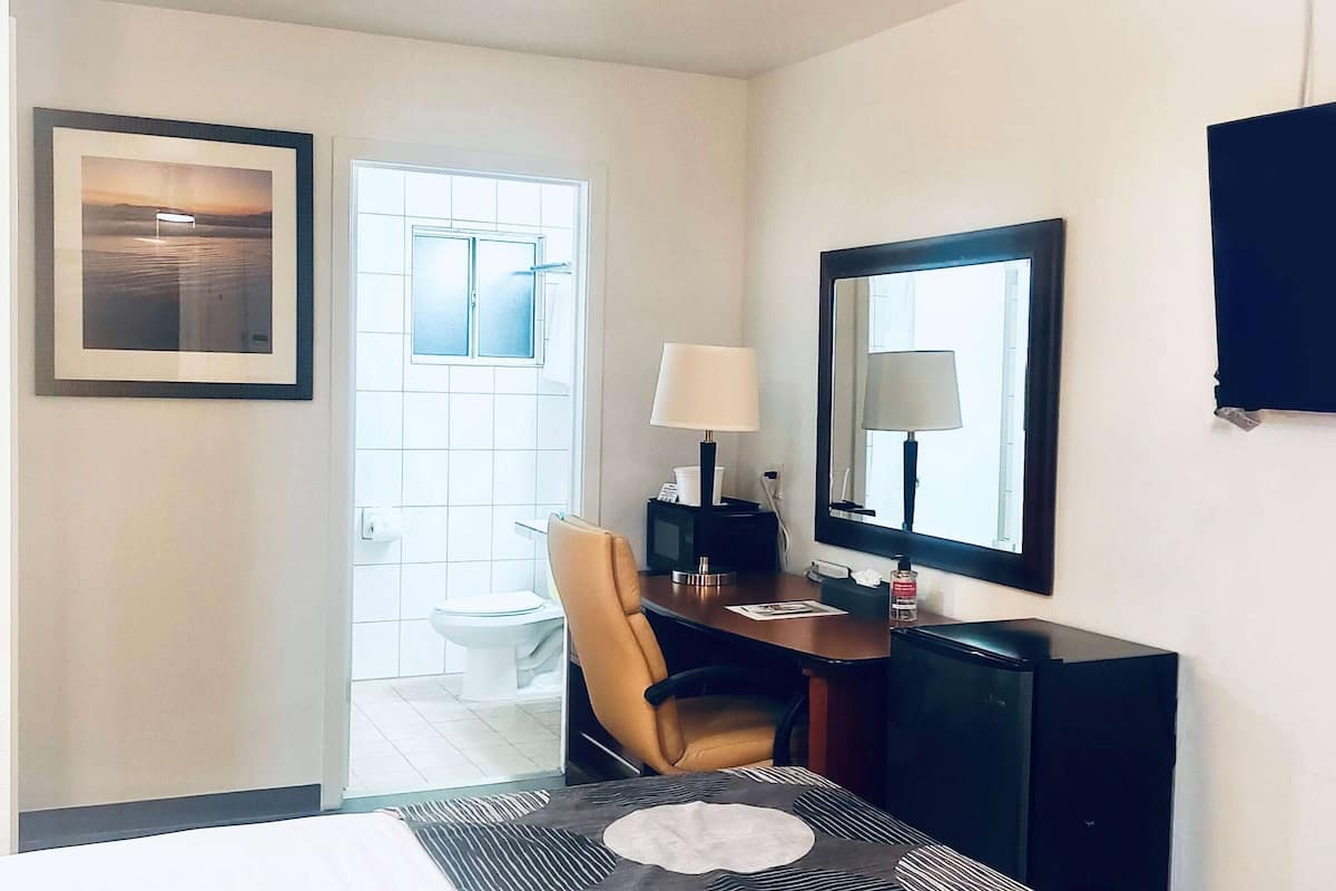 A hotel room with a bed, a desk with a chair, a television, a mirror, and a window with a view of a bathroom.