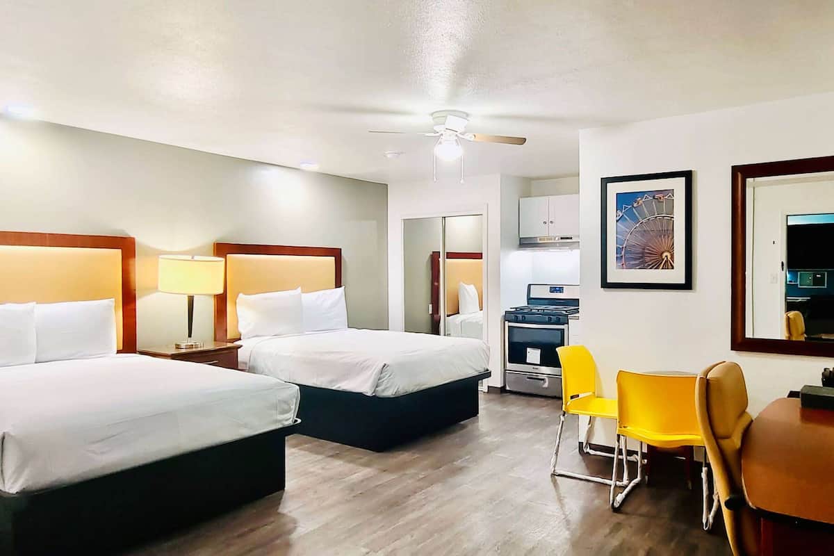 A hotel room with two beds, a desk, chairs, and a kitchenette.