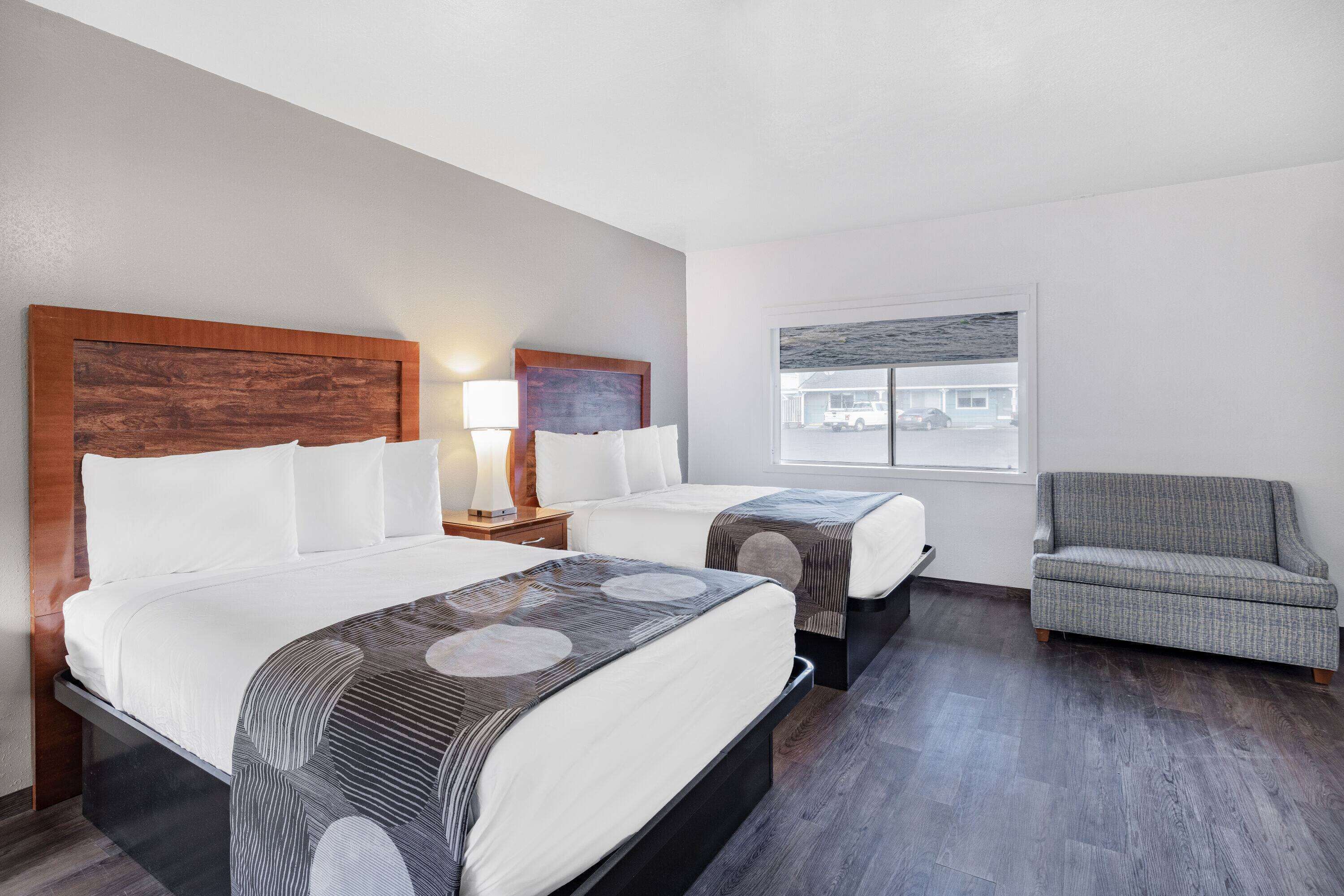 Une chambre d’hôtel avec deux lits, un fauteuil gris, une tête de lit en bois, une table de chevet avec une lampe et une fenêtre donnant sur un immeuble.