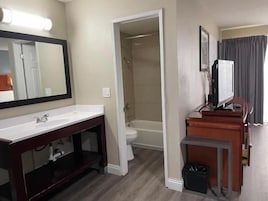 A hotel bathroom with a sink, mirror, shower, toilet, and a television on a wooden cabinet.