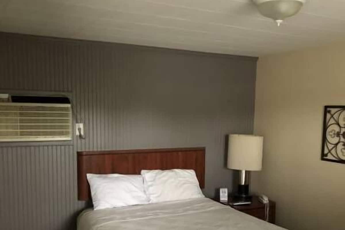 A neatly made bed with white linens, a wooden headboard, a bedside lamp, and a wall-mounted air conditioning unit.