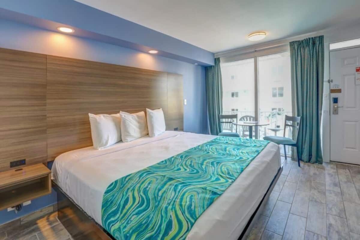 Une chambre d’hôtel avec un grand lit, une tête de lit en bois, un balcon avec une table et des chaises, et une vue sur la ville.