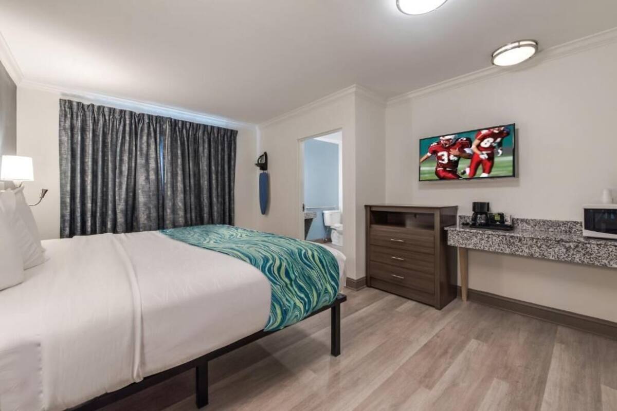 Une chambre d’hôtel avec un lit, un bureau équipé d’un ordinateur, une télévision et une salle de bain visible à travers une porte ouverte.