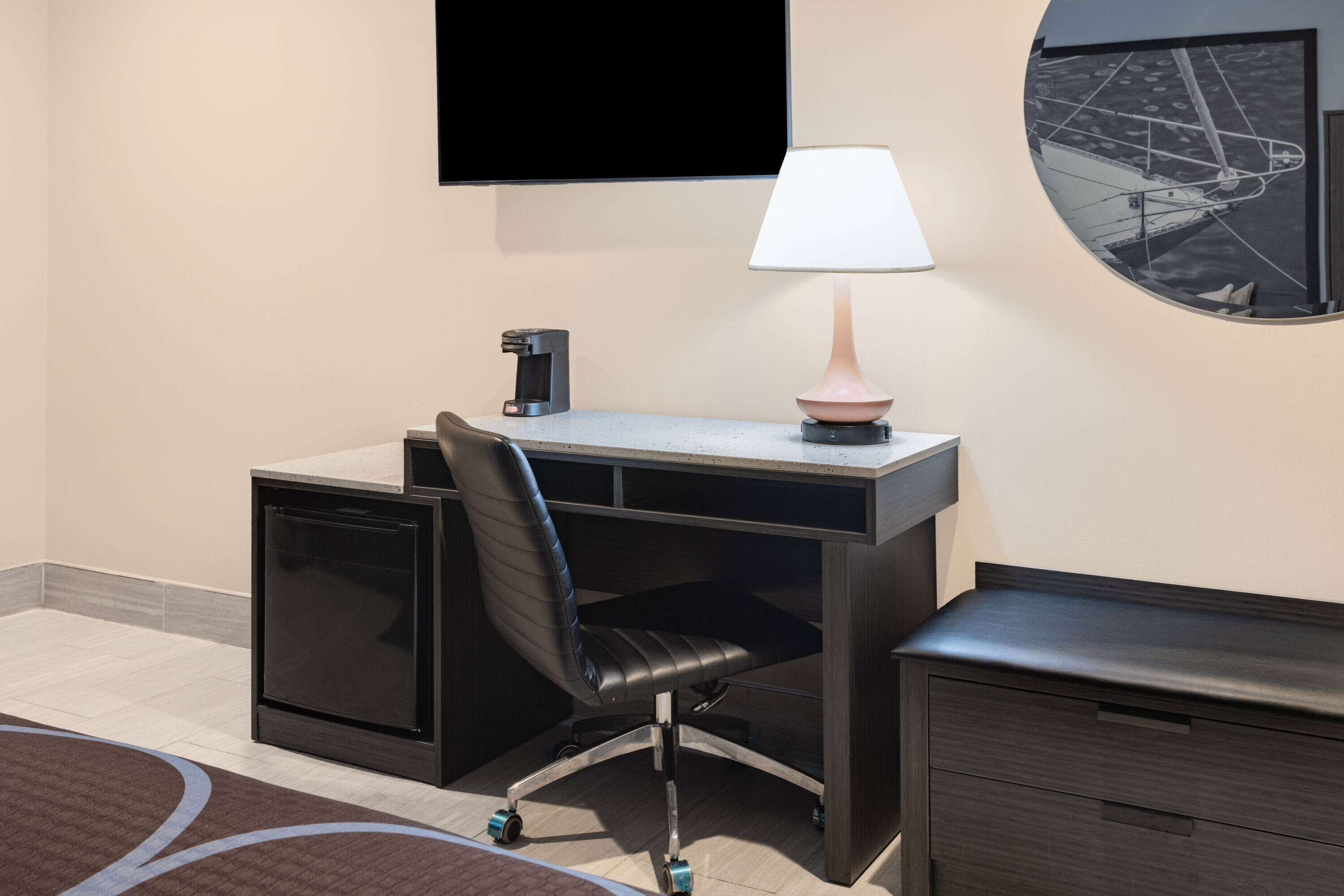 A hotel room with a desk, chair, TV, lamp, and a small cabinet.