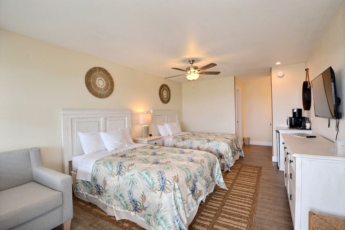 A hotel room with two beds, a ceiling fan, a desk with a TV, and a small seating area.