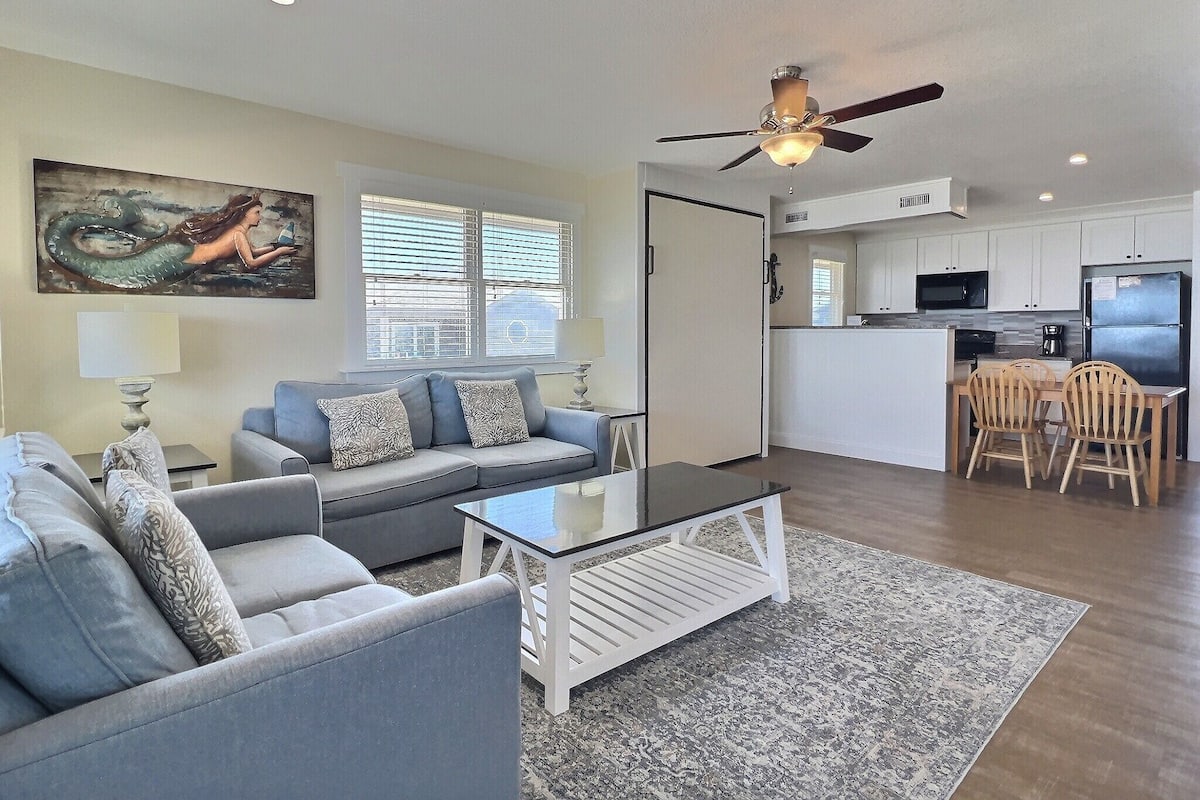 A modern living room with a grey sofa, a coffee table, and a dining area with wooden chairs.