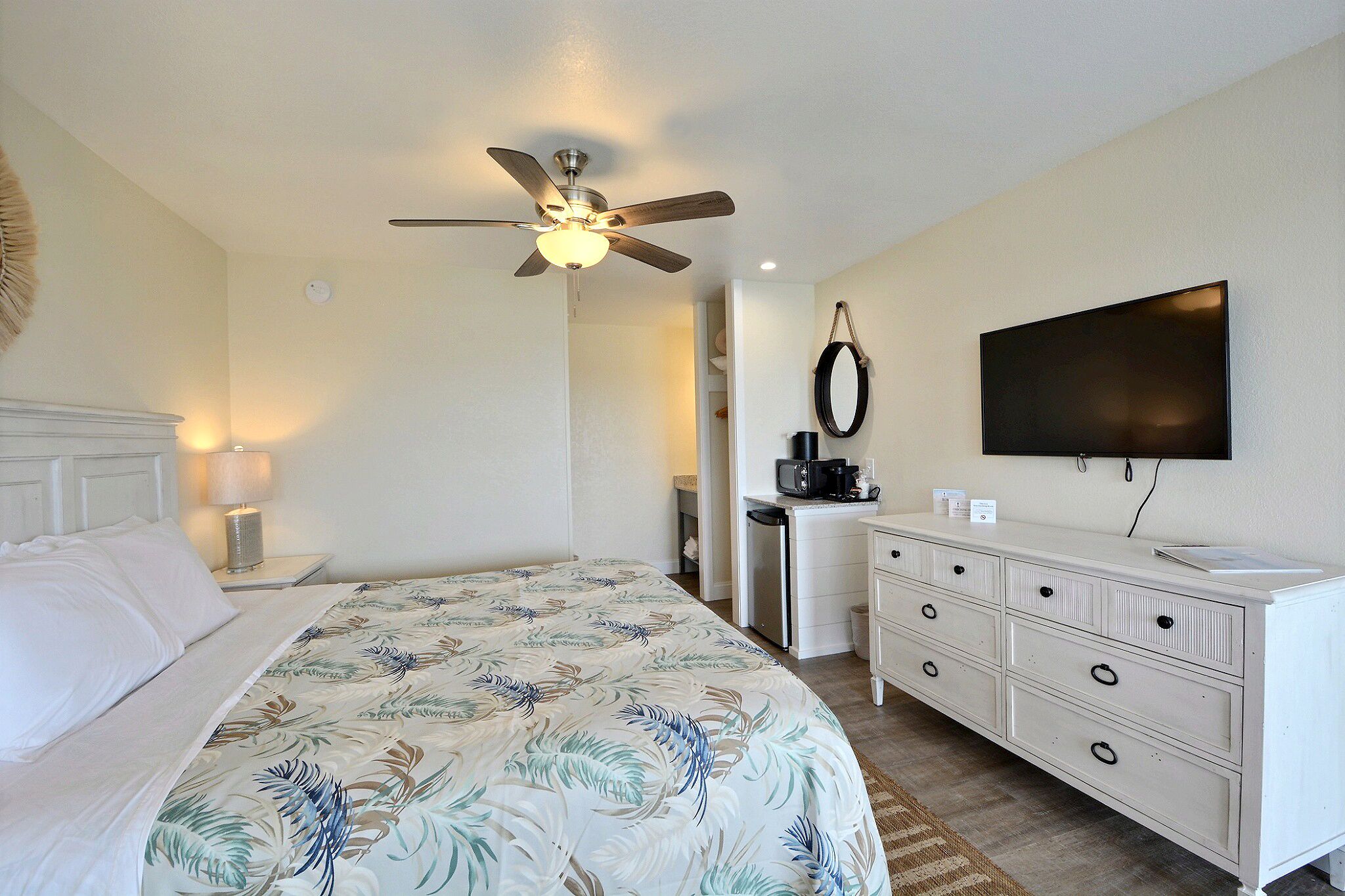 A hotel room with a bed, a ceiling fan, a TV, a dresser, and a desk with a coffee maker.