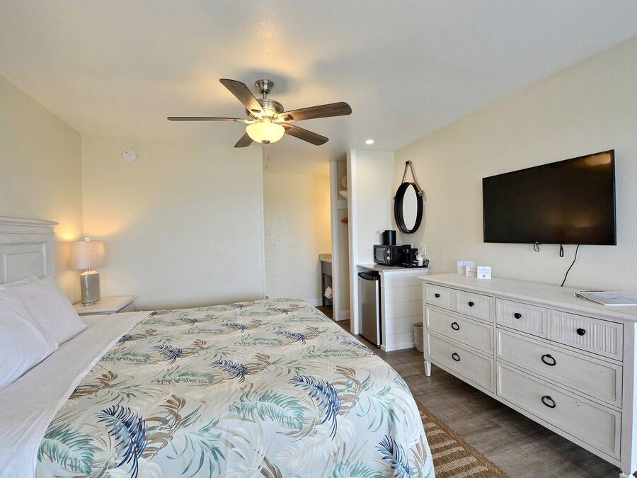 A hotel room with a bed, a ceiling fan, a TV, a dresser, and a desk with a coffee maker.