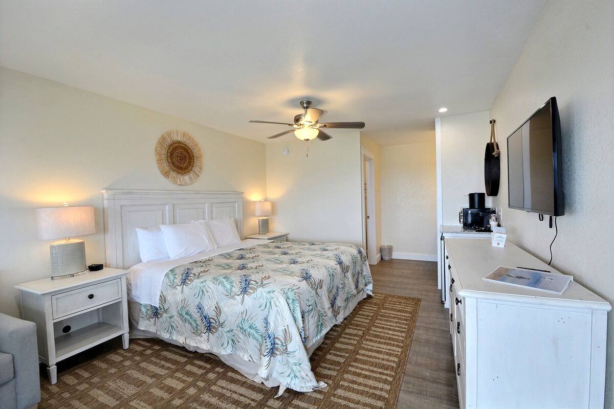A hotel room with a bed, nightstands, a desk, a ceiling fan, a television, and a wall-mounted coffee maker.