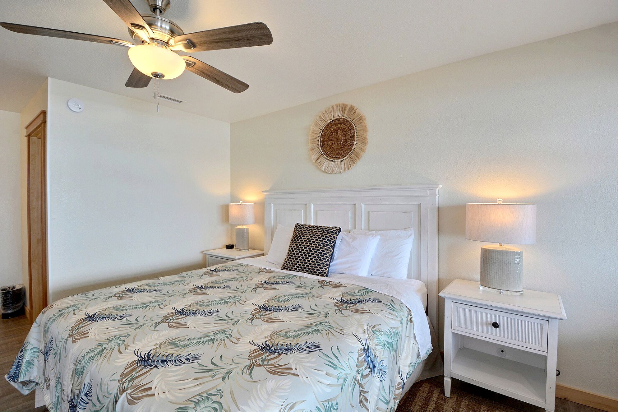 A bedroom with a bed, two bedside tables with lamps, a ceiling fan, a wall clock, and a decorative wall hanging.